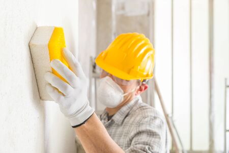 Plasterer or building wearing a hardhat and face mask for dust sanding and smoothing an interior wall during construction or renovation workの写真素材