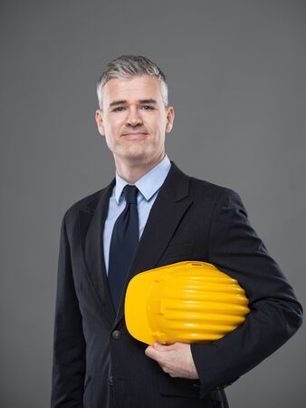 Architect or structural engineer carrying hardhat under his arms standing smiling at the camera over grey with copy spaceの写真素材