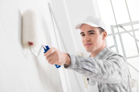 Friendly young male painter painting a white wall with a roller and smiling in a DIY, renovations or construction conceptの写真素材