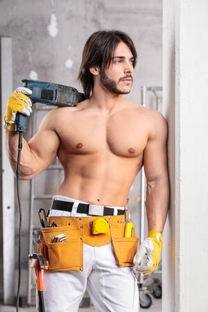 Handsome sexy muscular builder posing shirtless with a toll belt around his hips and power drill slung over his shoulder inside a room under constructionの写真素材