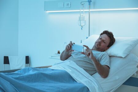 Adult sleepless patient lying in a hospital bed at night, he is watching video with his smartphoneの写真素材