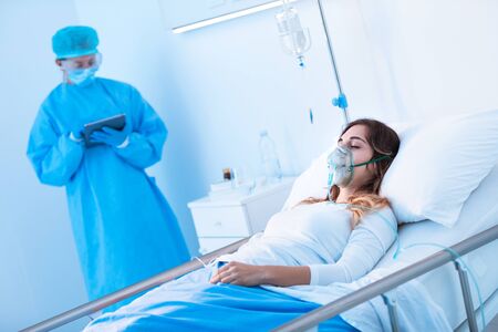 Nurse writing up notes on a Covid-19 patient lying in bed on the hospital ward wearing a positive pressure oxygen mask during the pandemicの写真素材