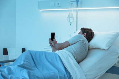 Sleepless worried patient lying in bed late at night at the hospital and chatting with his smartphoneの写真素材