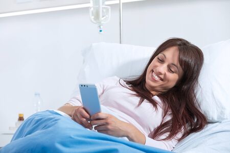 Smiling female patient lying in bed at the hospital and chatting with his smartphoneの写真素材