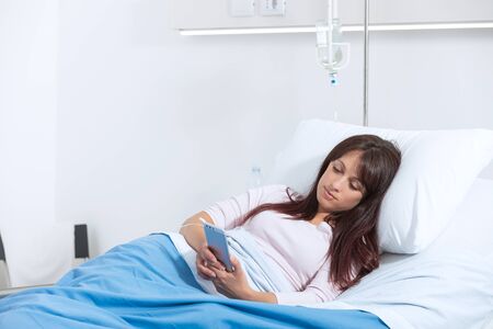 Sleepless female patient lying in bed at the hospital and chatting with his smartphoneの写真素材