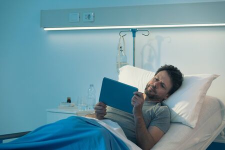 Adult sleepless patient lying in a hospital bed at night, he is watching movies with his digital tabletの写真素材