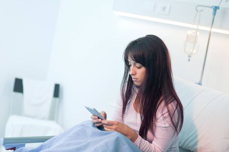 Sleepless female patient lying in bed at the hospital and chatting with her smartphoneの写真素材