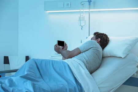 Adult sleepless patient lying in a hospital bed at night, he is watching video with his smartphoneの写真素材