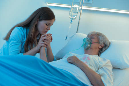 Worried daughter or granddaughter holding the hand of her mother in hospital as the elderly woman lies asleep or in a coma on a bed wearing a positive oxygen mask to assist breathingの写真素材