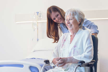 Carer or young daughter wheeling an elderly woman in a wheelchair bending down to smile at her indoors in a hospital room on a ward or old-age homeの写真素材