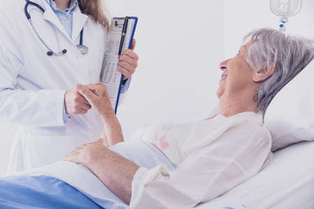 Smiling female doctor or nurse comforting an elderly woman patient in a bed reaching out to hold her hand during ward rounds at a hospitalの写真素材