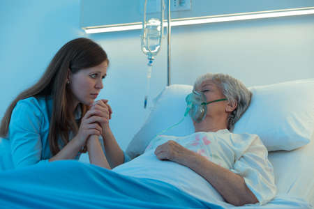 Worried daughter or granddaughter holding the hand of her mother in hospital as the elderly woman lies asleep or in a coma on a bed wearing a positive oxygen mask to assist breathingの写真素材
