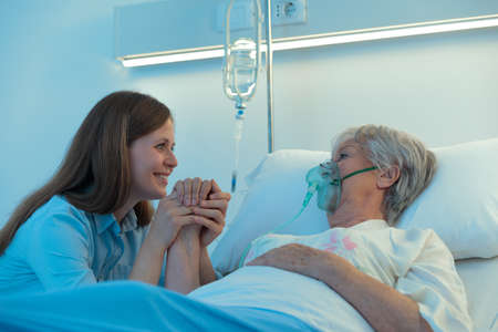 Young woman visiting her elderly mother or grandmother in hospital as she lies in a bed using a positive pressure oxygen mask to aid her breathing with a comforting positive smile as she clasps her handの写真素材