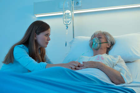 Worried daughter holding the hand of her mother in hospital as the elderly woman lies asleep or in a coma on a bed wearing a positive oxygen mask to assist breathingの写真素材