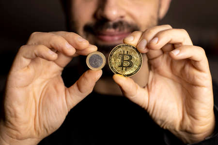 Caucasian hands shows one Bitcoin coin and one Euro Coin. New finance concept. Comparison between currencies, fiat and crypto. Two hands. Man smiles in the backgroundの写真素材