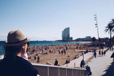 Young and stylish woman standing on the seafront next to the sandy beach and looking tourists taking rest along Barceloneta beachの写真素材
