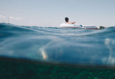 half underwater view of a young boy on rubber dinghy floating on the sea of Sardinia - Italy.の写真素材