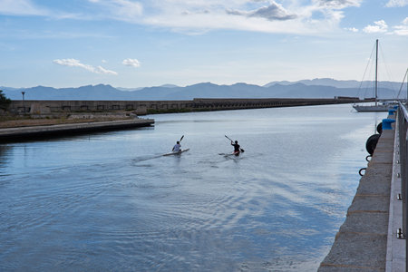 Two kayakers paddling on calm water with mountains and harbor in the background in Cagliari,...