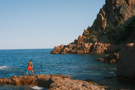 A solitary figure stands on a rocky shore, gazing at the serene blue waters. The jagged cliffs rise majestically in the background, capturing the essence of adventure and tranquility in nature.の写真素材