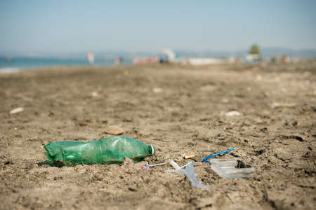 Green, plastic bottle, cotton swabs, cap, box and cigarettes left on a sandy beach.の写真素材