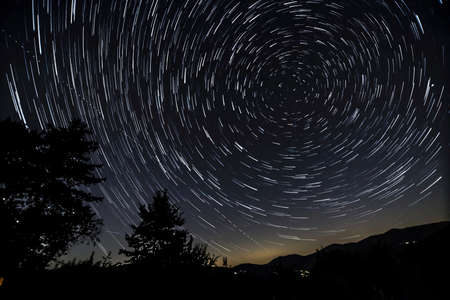 Star trail photograph around the North Polar star with hills, trees and house lights.の写真素材