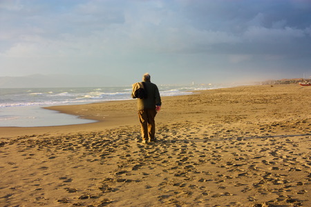 an elderly man wanders on the beach. maybe he's lost and he can not find his way home anymore. he wanders in his loneliness, in his melancholy. mental illness, perhaps parkinson's or alzheimer's.の写真素材