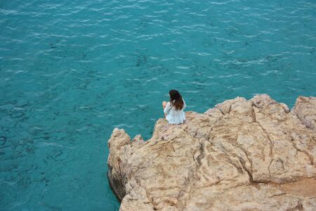 figure from the back, in the distance, of a girl unrecognizable who scrutinizes the sea, the infinite, sitting alone on a rocky cliff on the ocean water in a white dress in ibiza, spainの写真素材