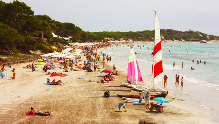 sandy beach crowded with ses salines in ibiza and bathers by the sea in summer on the shoreline in spainのeditorial素材