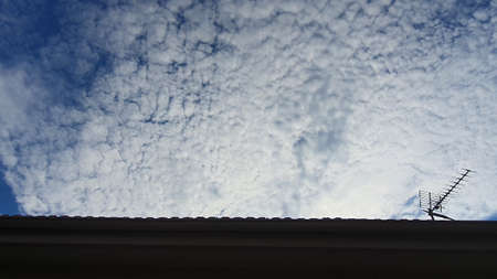 Shot of a cloudy daytime sky with a backlit house top and a TV antenna.