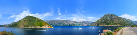 A panorama of the black mountains surrounding the bright blue gulf of the bay, cutting white yachts. The giant mountain, at the foot of which small houses have accumulated, is reflected in the brightly blue surface of the bay and the yacht.の写真素材