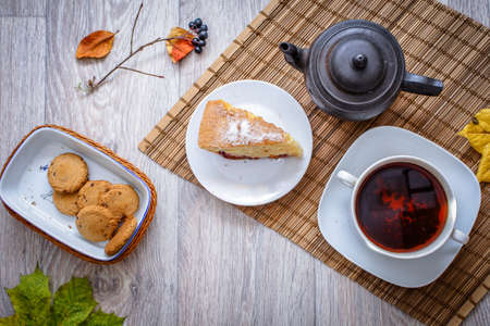 An old teapot on a straw bedding with a cup of hot tea a slice of plum sponge cake and chocolate...