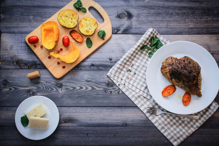 A piece of meat, sprinkled with spices, baked with baked carrot slices on a white plate, fork, linen checkered napkin, next to the baked slices of pumpkin, potatoes, spices and cherry tomatoes.の写真素材