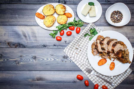 Composition of sliced meat baked in spices, carrot slices baked on a white plate on a checkered linen napkin, surrounded by cherry tomatoes, carrots baked on a plate next to potatoes; bread plate and bowl of peppersの写真素材