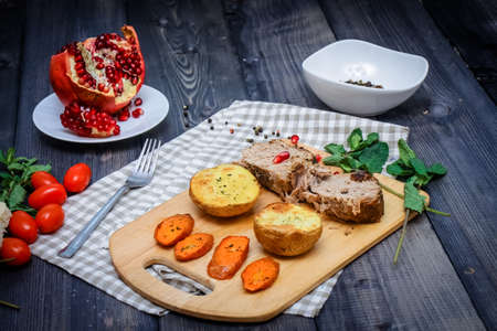 On the cutting board are two slices of baked meat, two slices of potatoes and baked carrot slices, a checkered napkin of flax, in a bowl are multi-colored peas of pepper and a cut pomegranate on a plate against the graphite tabletopの写真素材