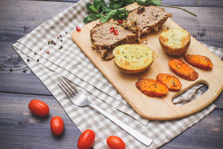 On the cutting board are two slices of baked meat, two slices of potatoes and baked carrot slices, a...
