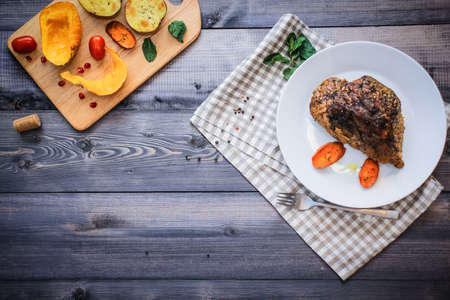 A piece of meat, sprinkled with spices, baked with baked carrot slices on a white plate, fork, linen checkered napkin, next to the baked slices of pumpkin, potatoes, spices and cherry tomatoes.の写真素材