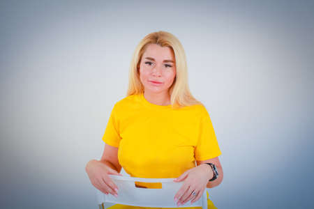 White to light blue gradient background. A girl in a yellow t-shirt looks at the camera and sits on a chair, leaning back against a chair.の写真素材