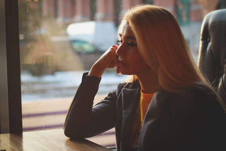 Cafe By the window daylight. A blonde girl is sitting at a table in a cafe propping up her cheek.