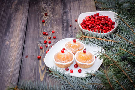 A light wooden table top with a plate of freshly baked muffins decorated with red berries sprinkled with white powder and a plate of red berries with spruce twigs.の写真素材