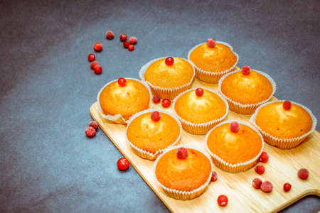 On gray craft paper there is a wooden cutting board on which are freshly baked muffins decorated with red berries scattered on paper.の写真素材