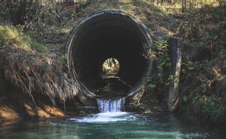 The black hole of the pipe, from which flows a blue stream, flowing into the lake.Autumn forest landscape. The colors of autumn.の写真素材
