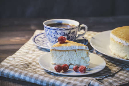 On a dark tabletop, on a light checkered linen napkin on a white saucer, a piece of cheesecake cake...