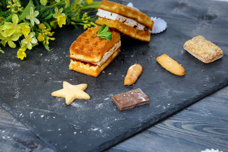 Near the spread of snow little meringue On graphite colors of flat stone meringue tiles there are shaped cookies, of different kinds, peaks of gentle beze, chocolate tiles, large Vienna waffles create the center of the composition against the background of a light yellow bouquet of field colors.の写真素材