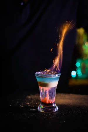 The fire scatters into sparks. In the glass, the cocktail is brown-red, the top is white and the top is emerald, which is set on fire, the layers are not mixed.の写真素材