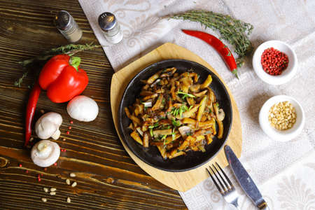 In a frying pan, fried potatoes frame pieces of meat stewed in sauce, cutlery, red bell peppers, champignons, sauce, hot peppers, spices, rosemary branch are on the table. Fried potatoes are served in a frying panの写真素材