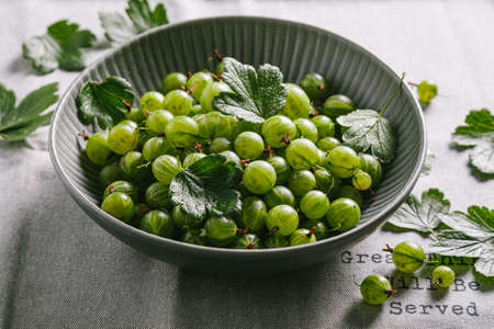 Green gooseberries with leaves in a gray ceramic plate on a gray kitchen towel. Close up.の写真素材