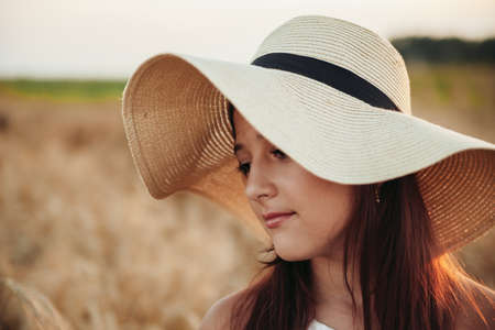 Happy girl in a hat stands in a field of ripe wheat in the sunset light. Selective focus. Sunset sun. Walking in the wheat field.の写真素材