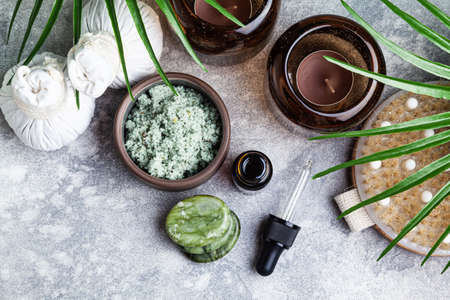 Set of natural skin care products and palm leaves on a gray background. Massage stones, essential oil, herbal compress stamps and scrub. Spa treatment concept.の写真素材