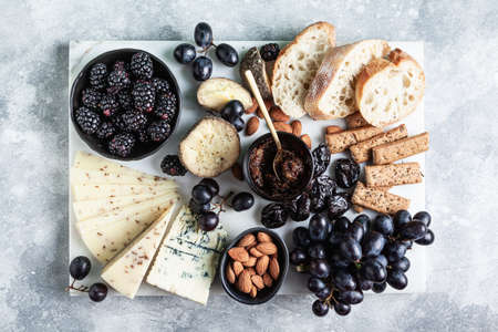 Different types of cheese with fruits, berries, nuts and bread on a white marble board on a gray...