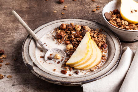 Chocolate granola with almond milk, pear and honey in a ceramic plate on a brown background. Healthy breakfast or snack.の写真素材
